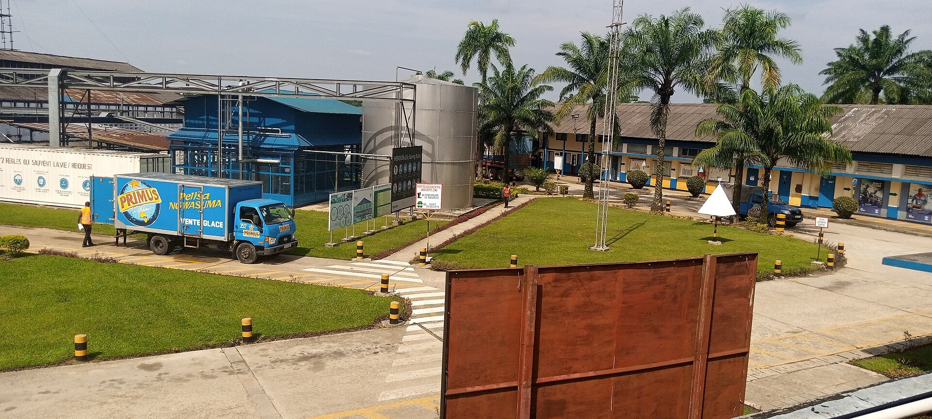 A brewery yard in Kisangani with a blue Primus delivery truck, tanks, palm trees and low plant buildings, DR Congo