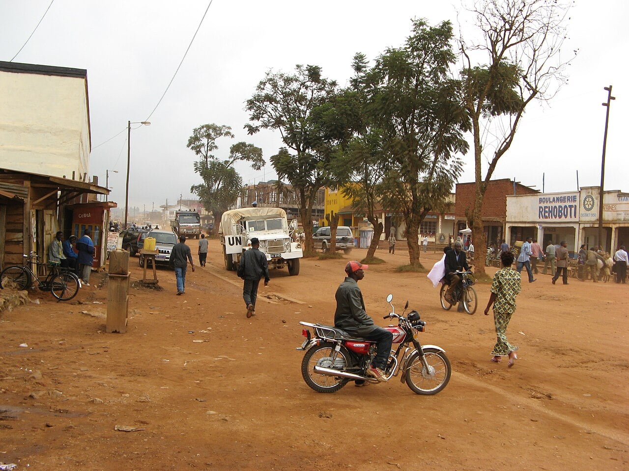 Motorcycle taxi rider, pedestrians and a United Nations truck on a dusty main street lined with shops in Butembo, DR Congo
