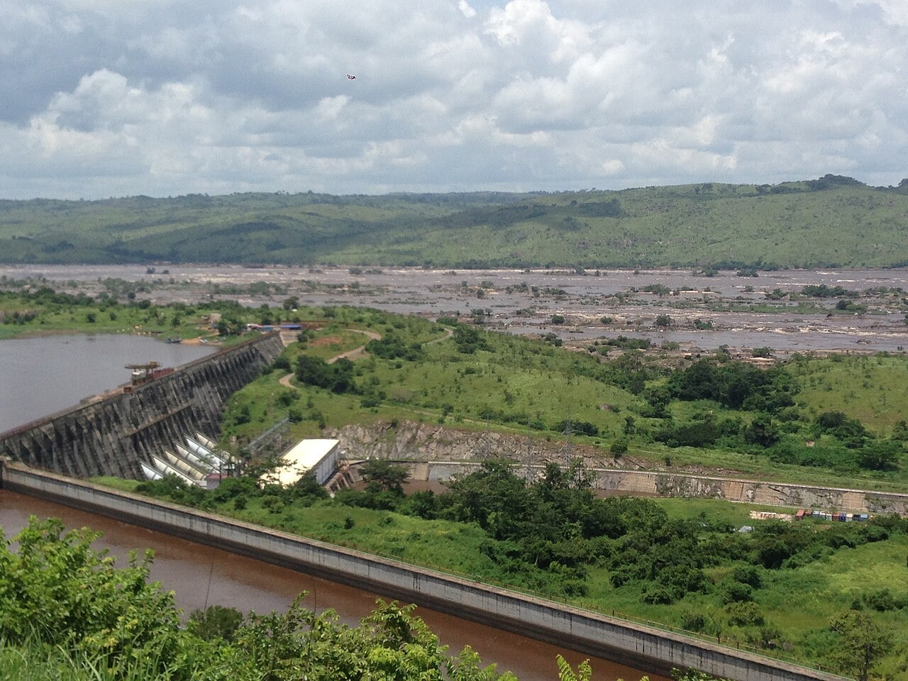A concrete dam with spillway gates holds back a reservoir beside the wide, rock-strewn rapids of the Congo River, with green hills behind