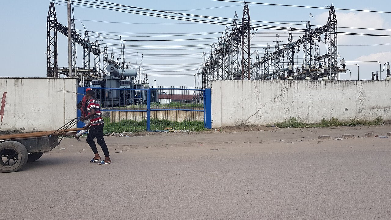 A man pulls a handcart past a walled SNEL electricity substation with transformers and pylons in Limete, Kinshasa