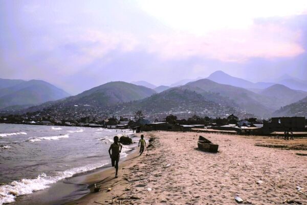 Boys run along the sandy Lake Tanganyika shore at Uvira, DR Congo, with wooden boat and hills behind