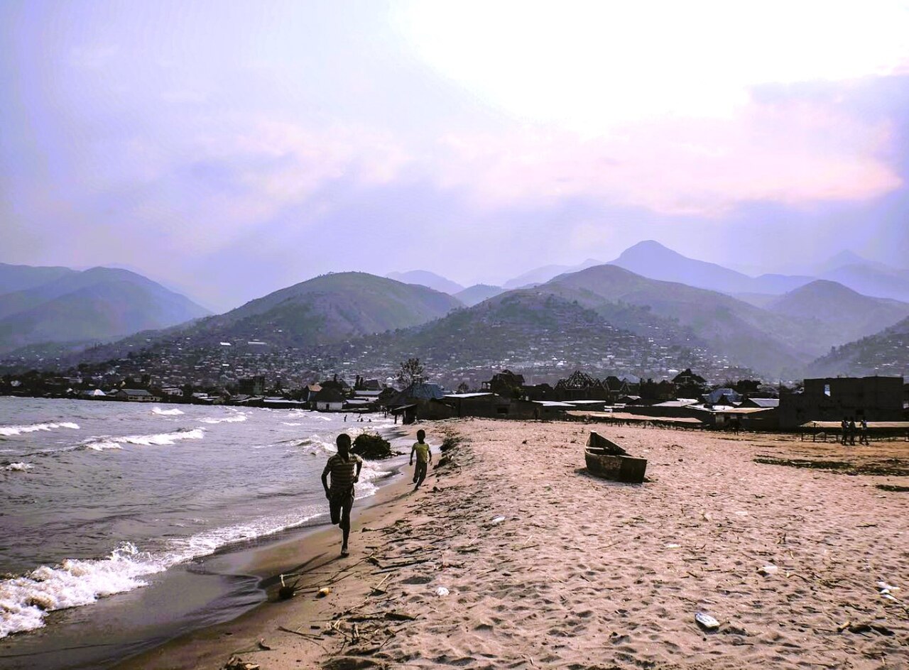 Boys run along the sandy Lake Tanganyika shore at Uvira, DR Congo, with wooden boat and hills behind