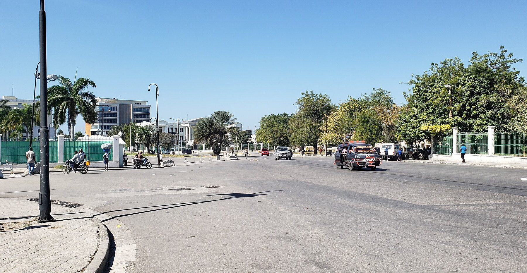 A wide sunny street at the Champ de Mars in Port-au-Prince, with palm trees, green railings, a minibus and motorcycles