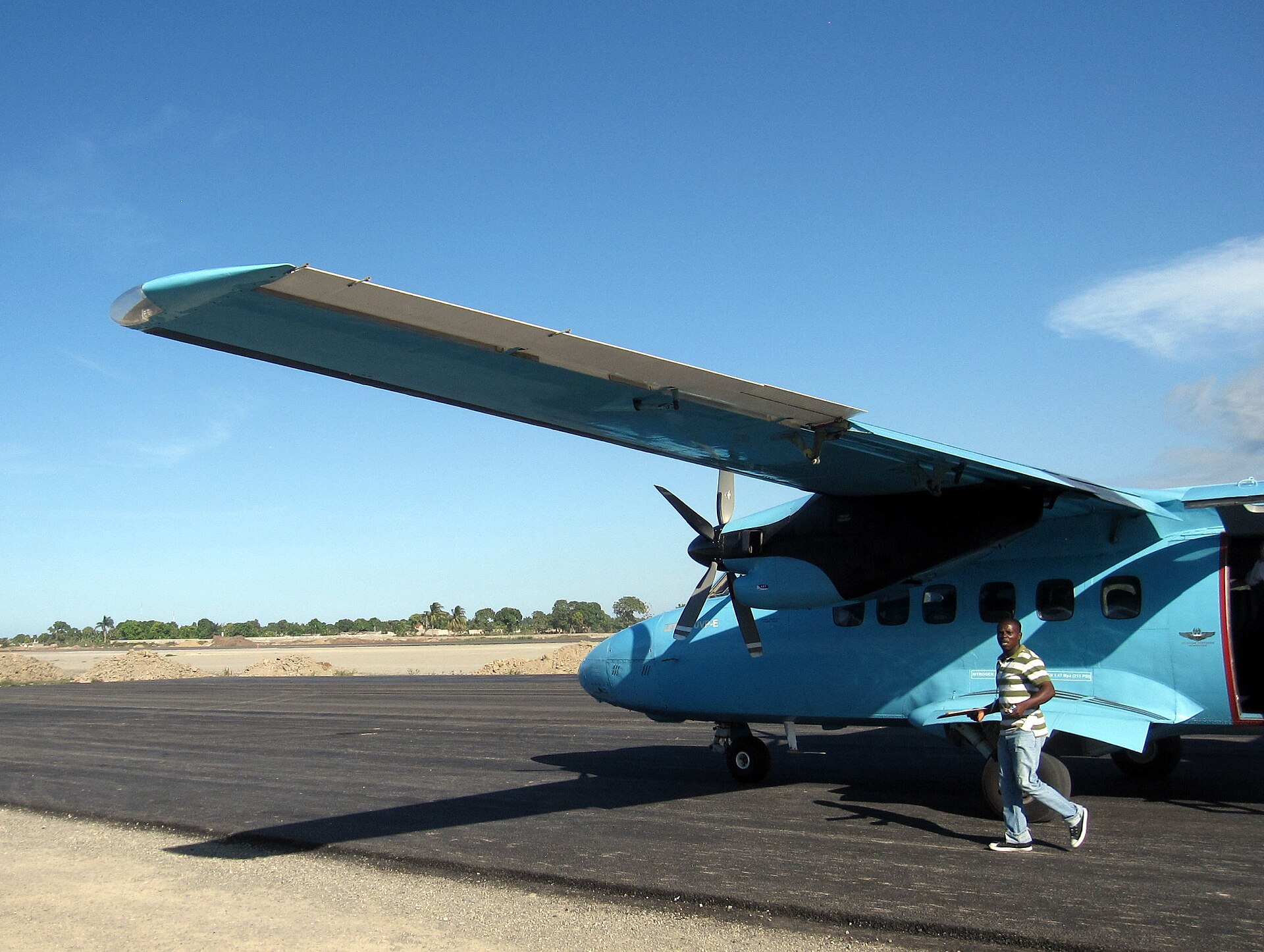 A small blue twin-propeller passenger plane on the apron at Cap-Haitien airport, with a man walking beside it