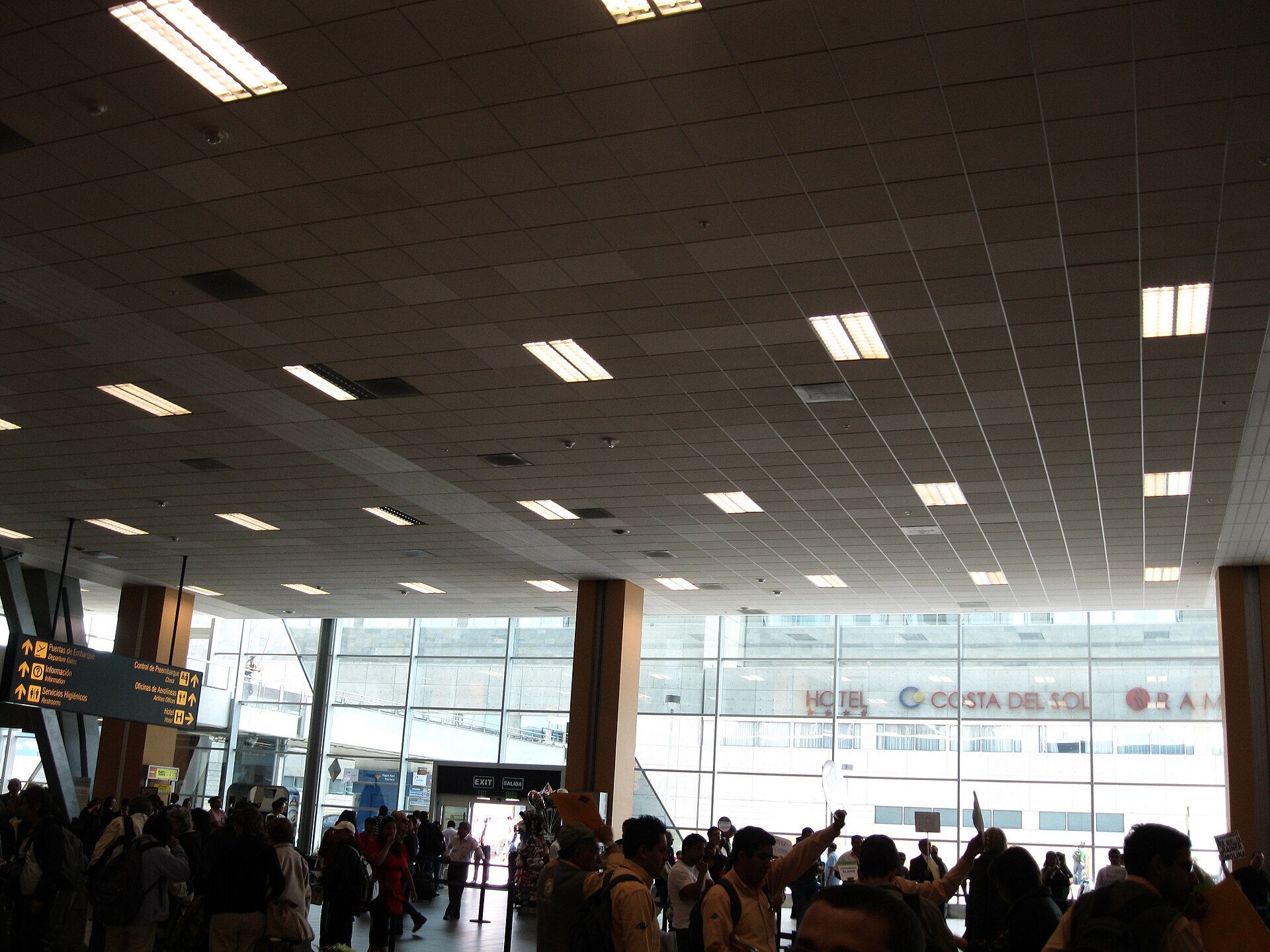 A crowded airport arrivals hall with a tiled ceiling, glass frontage and people holding up name signs