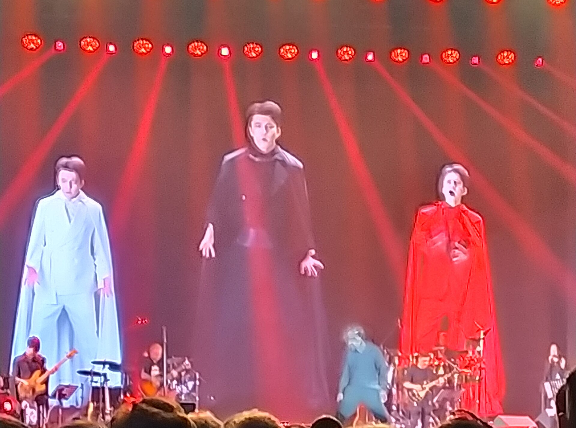Concert stage under red lights with three giant video images of Dimash Qudaibergen above his band, seen over the heads of the audience