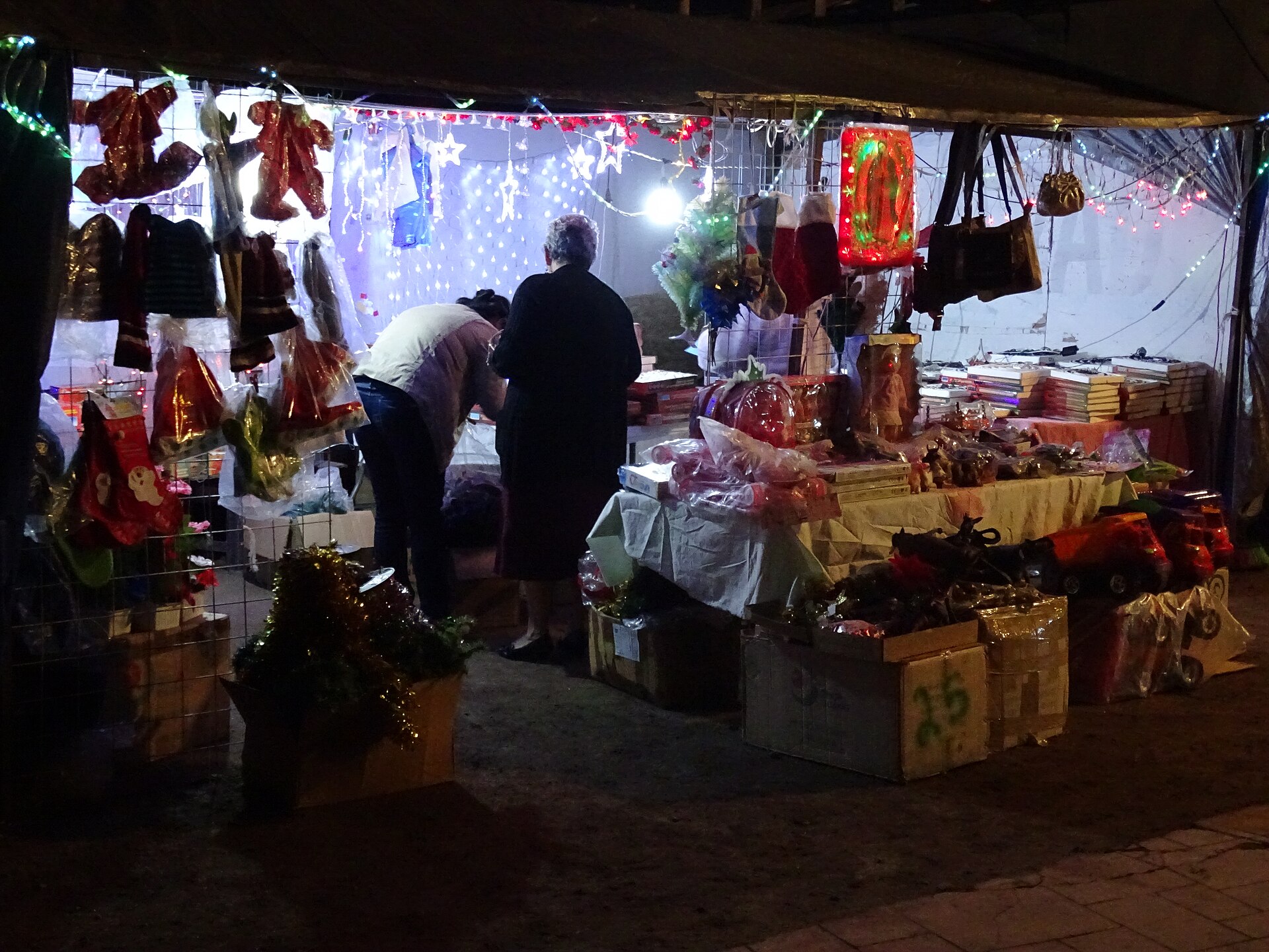Two people browse a lit night stall hung with Christmas stockings, decorations and toys