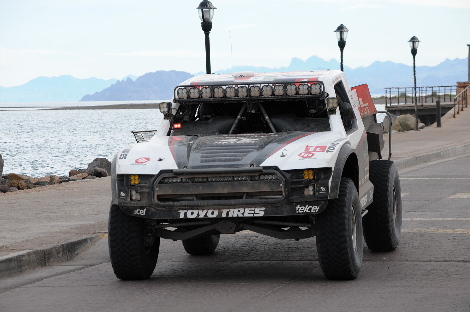White desert racing truck with a roof light bar and large off-road tyres on a seafront promenade, with sea and mountains behind