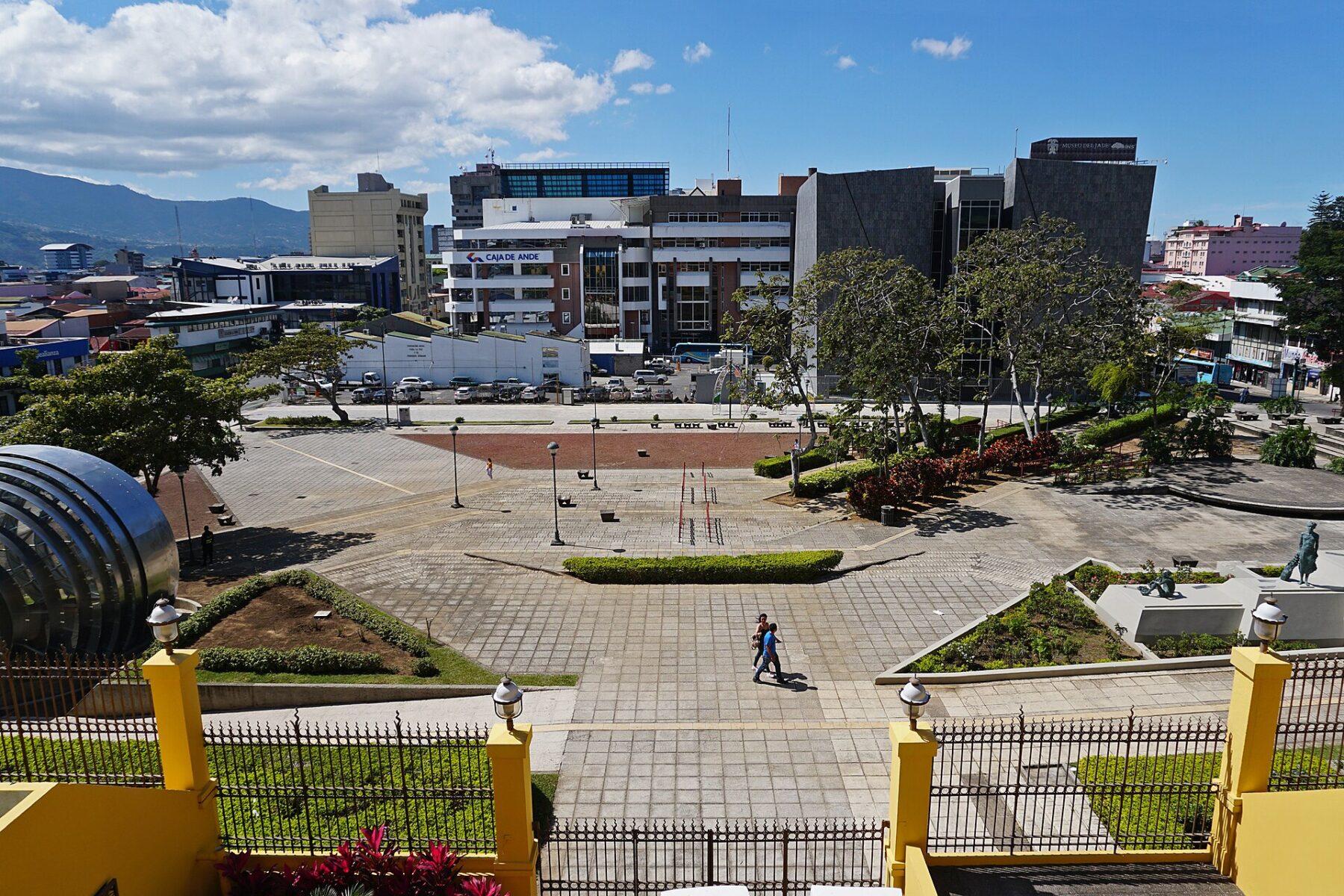 A wide paved square with lawns, flower beds and lamp posts, a curved metal dome on the left and office buildings and green hills behind