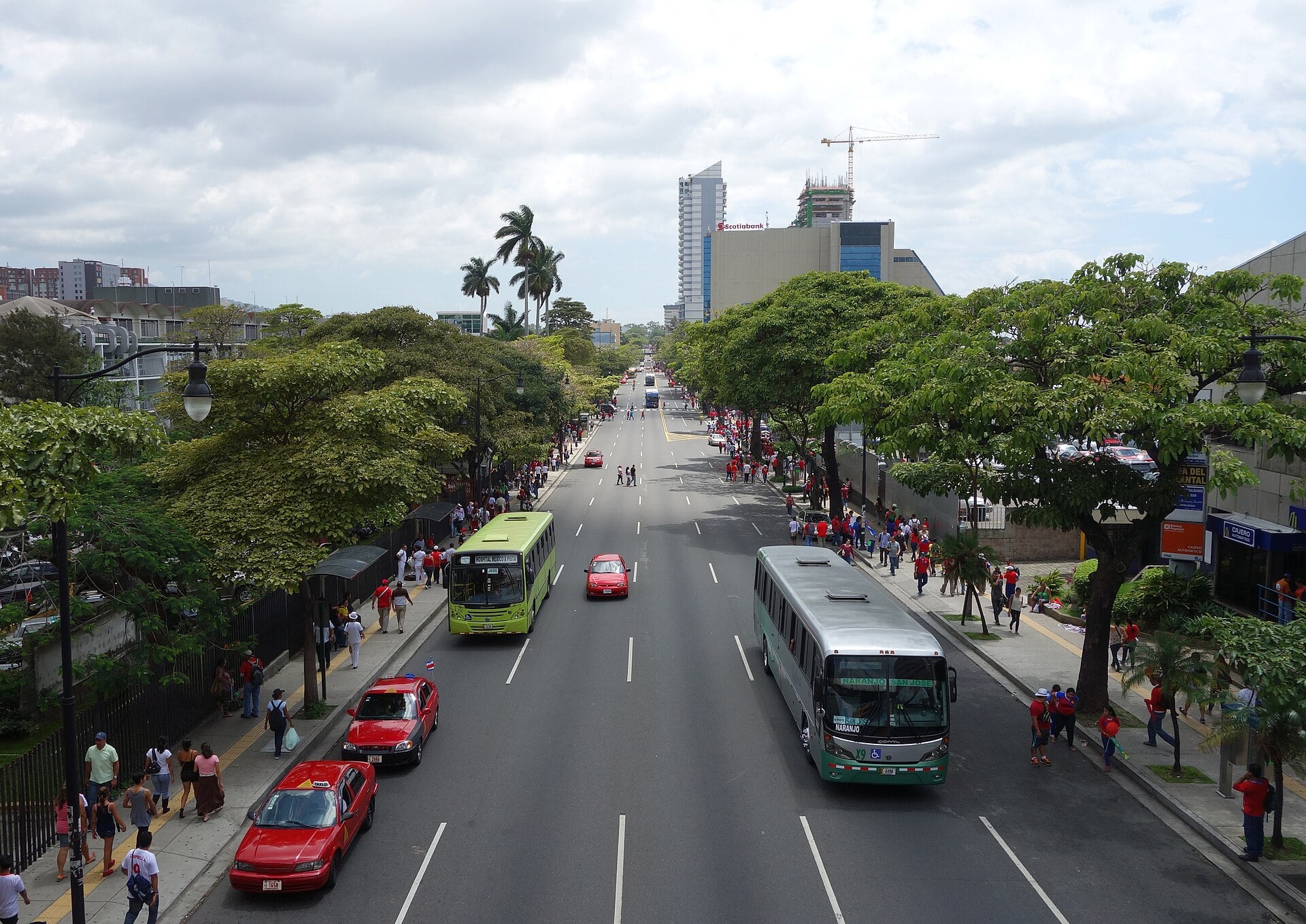 A wide tree-lined avenue with buses, red taxis and people on the pavements, high-rise towers and a construction crane in the distance