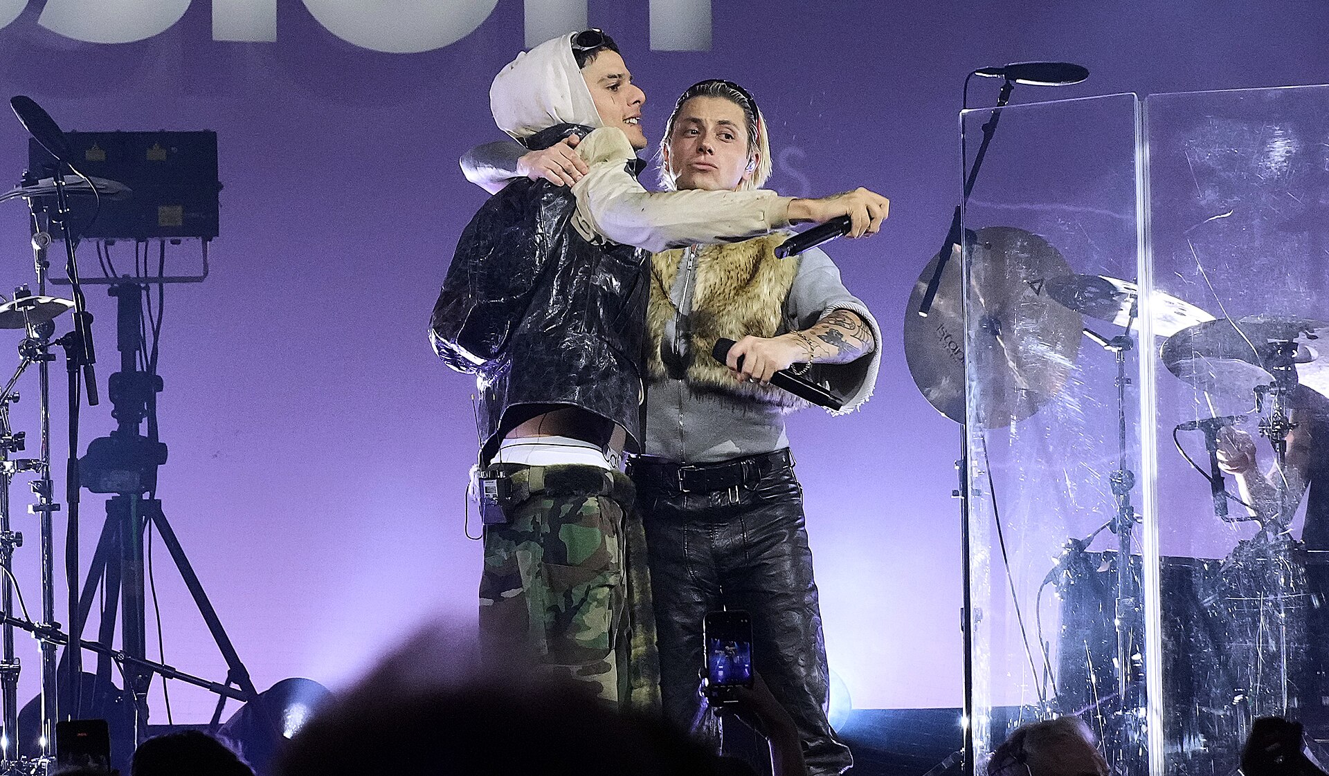 CA7RIEL and Paco Amoroso embracing on stage with microphones in hand, a drum kit behind a clear screen to the right