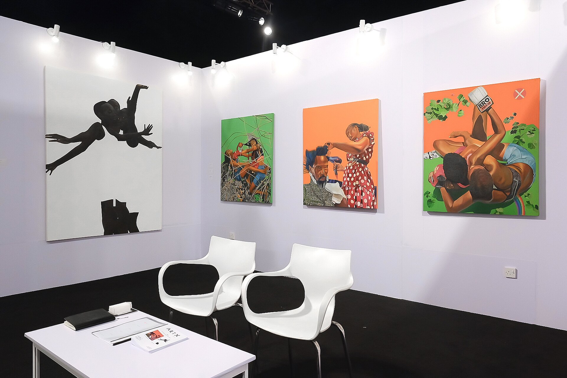 White-walled gallery booth with four paintings, two white chairs and a small desk