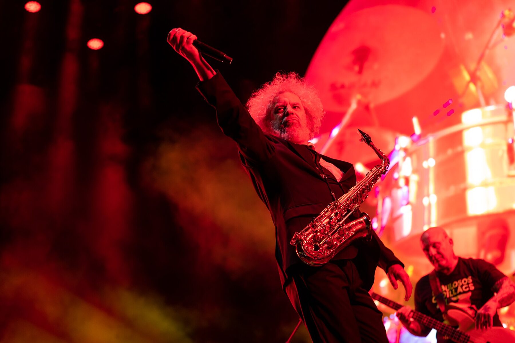 A curly-haired saxophonist raising a microphone on a red-lit stage, with a bassist in a Fabulosos Cadillacs T-shirt behind him