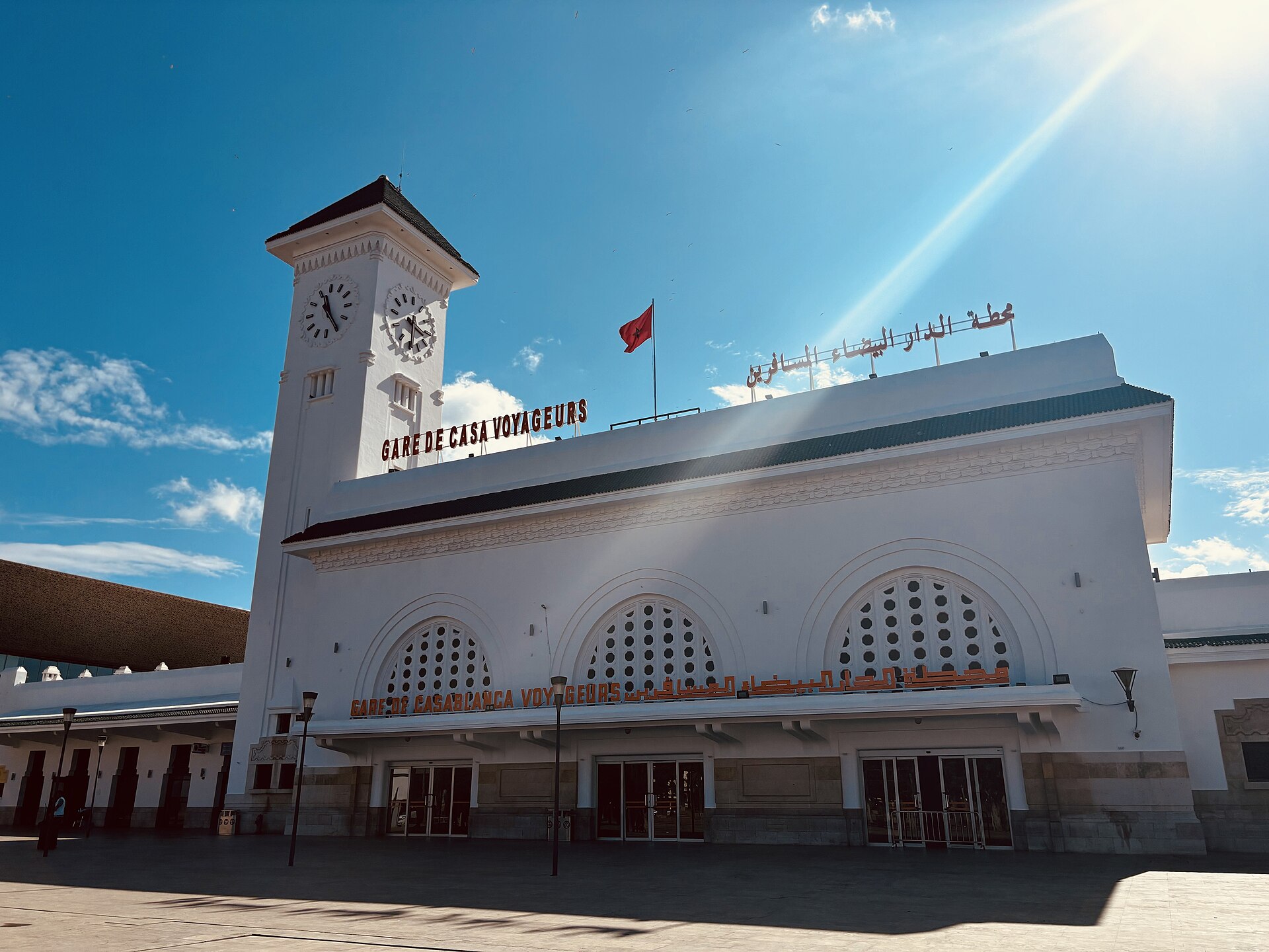 The white facade and clock tower of Casa Voyageurs railway station in Casablanca with a Moroccan flag on the roof