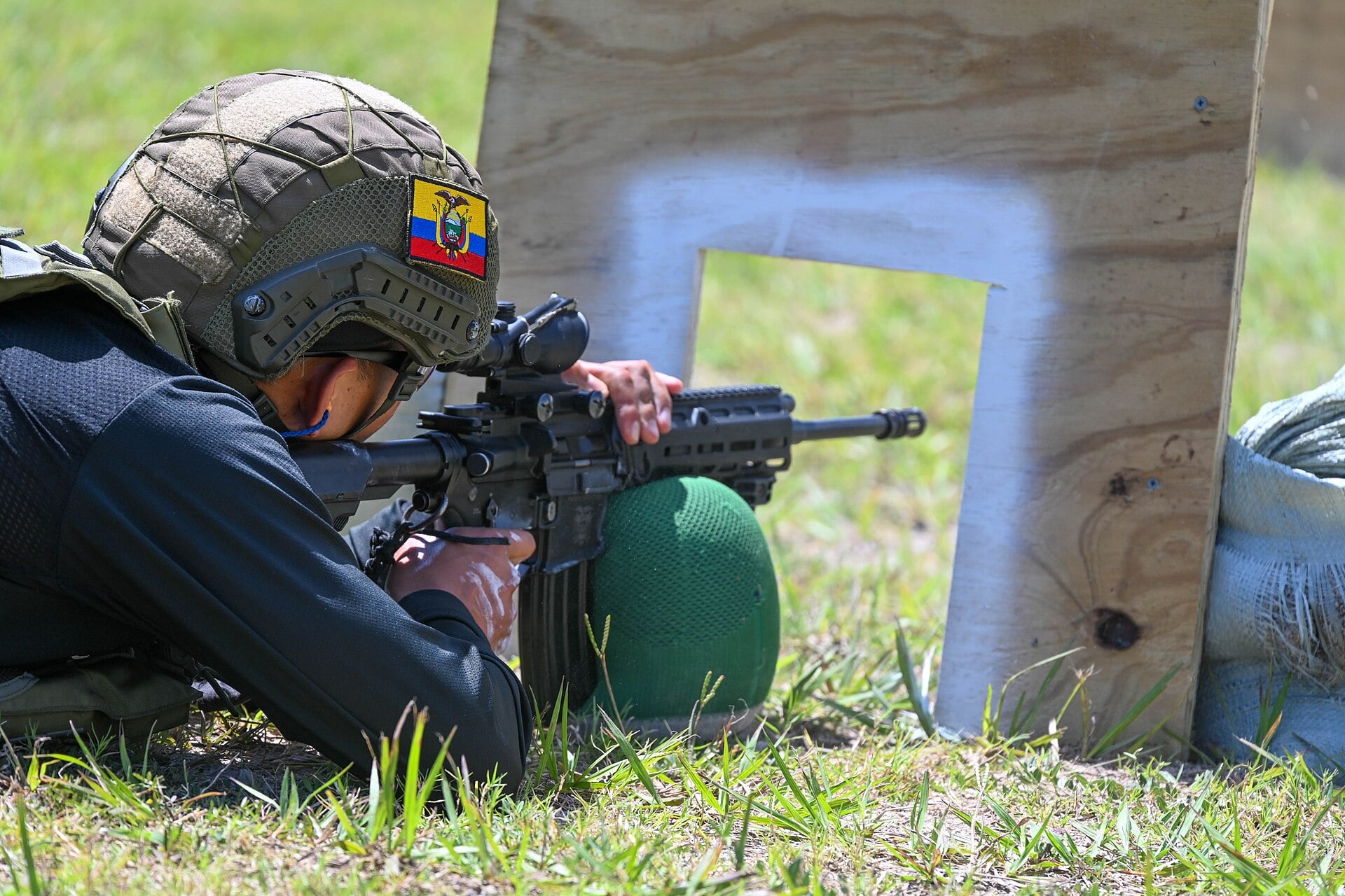 Ecuadorian soldier with a flag patch on his helmet lying on grass and aiming a rifle with a scope at a target board