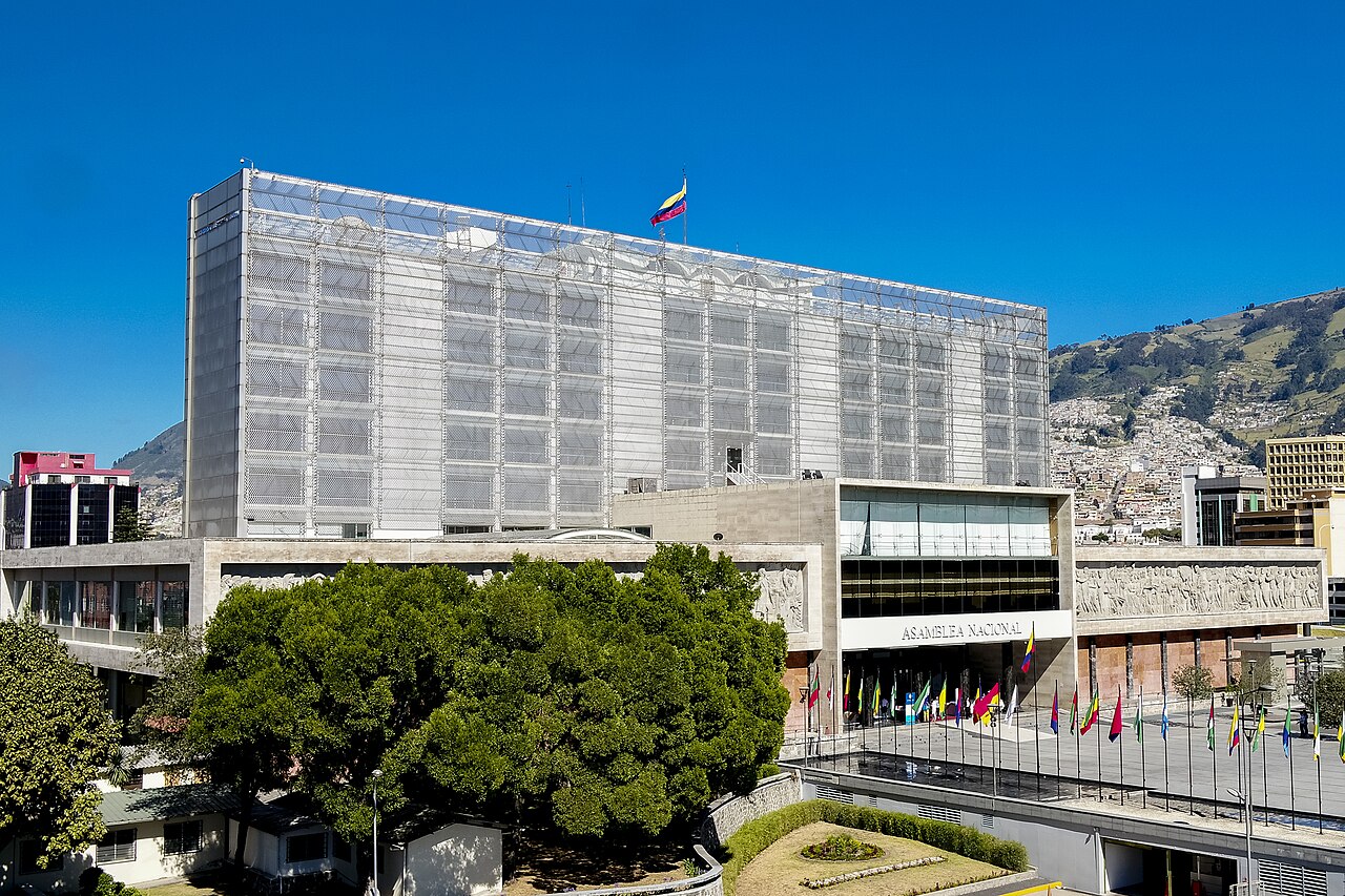 The National Assembly building in Quito, a tall white tower behind a long low block with flags in front and green hills behind