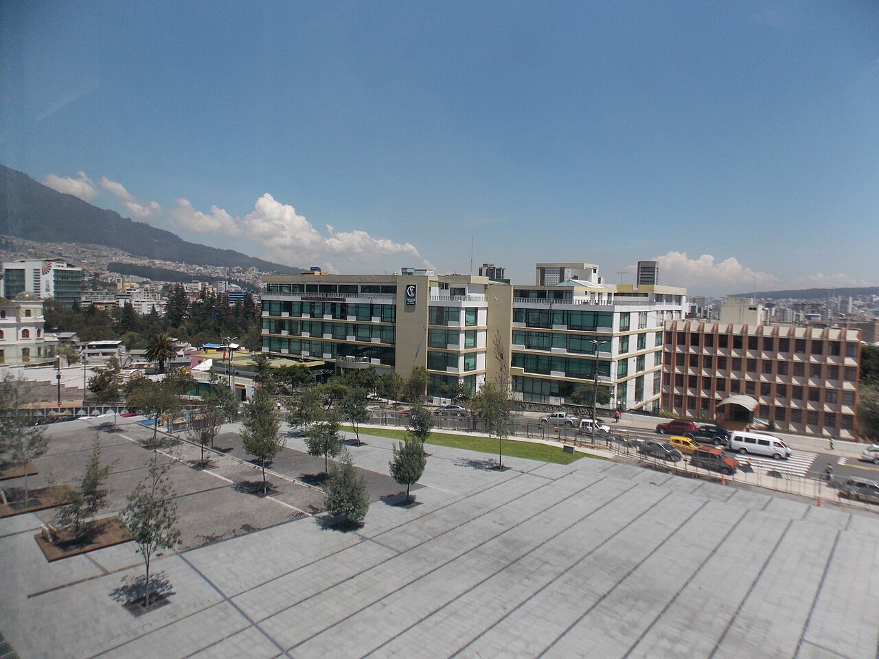 Paved entrance plaza with young trees in front of a modern office building marked with a C logo, with Quito hillsides behind