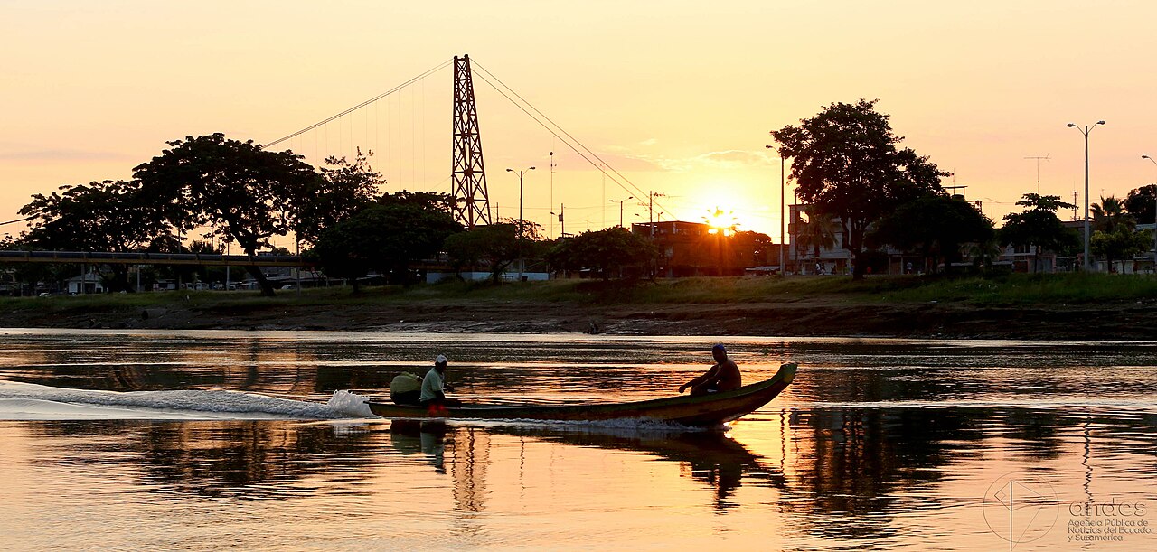 Two men ride a long wooden motorboat on the Babahoyo river at sunset, with a suspension bridge tower and trees on the bank behind
