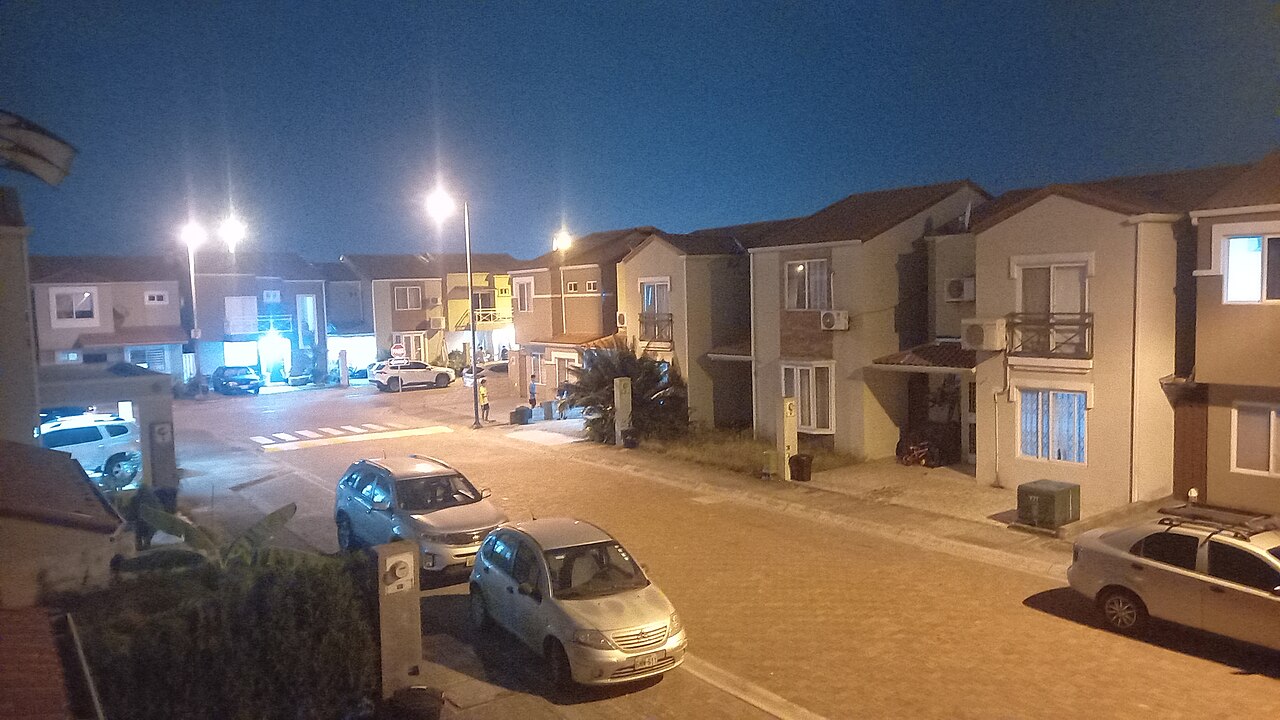 A residential street of two-storey houses in Guayaquil at night, lit by street lamps, with parked cars outside