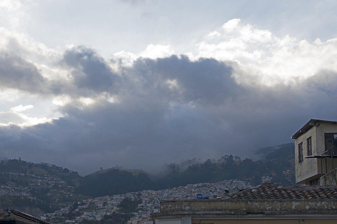 Hillside neighbourhoods of Quito under heavy grey clouds, seen over tiled rooftops in the historic centre