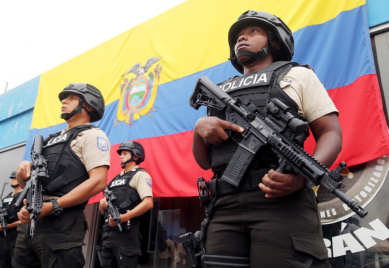 Helmeted police officers in body armour marked Policía hold rifles in front of a large Ecuadorian flag