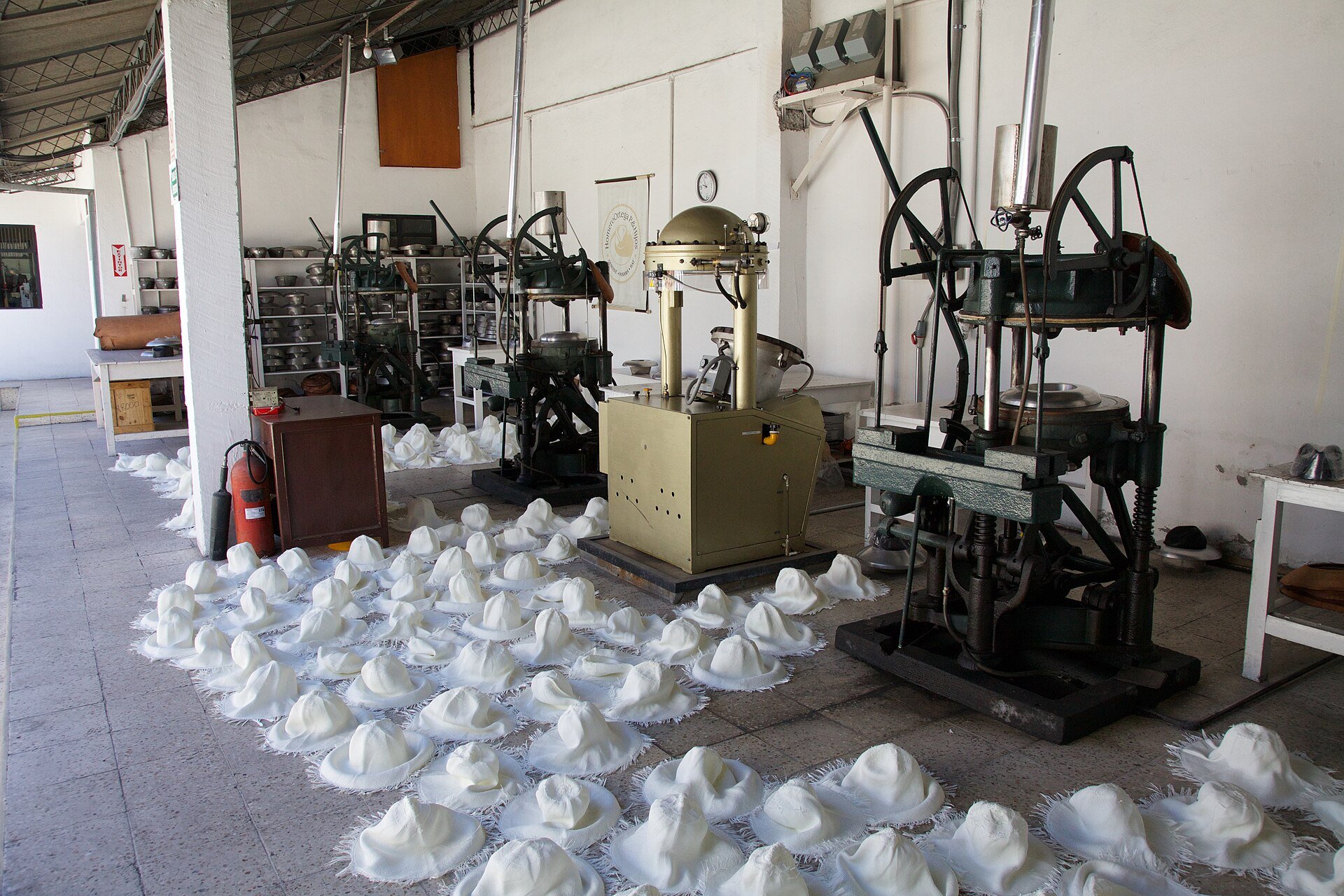 Rows of white woven toquilla straw hats on the floor of a hat factory in Cuenca, Ecuador, beside shaping machines