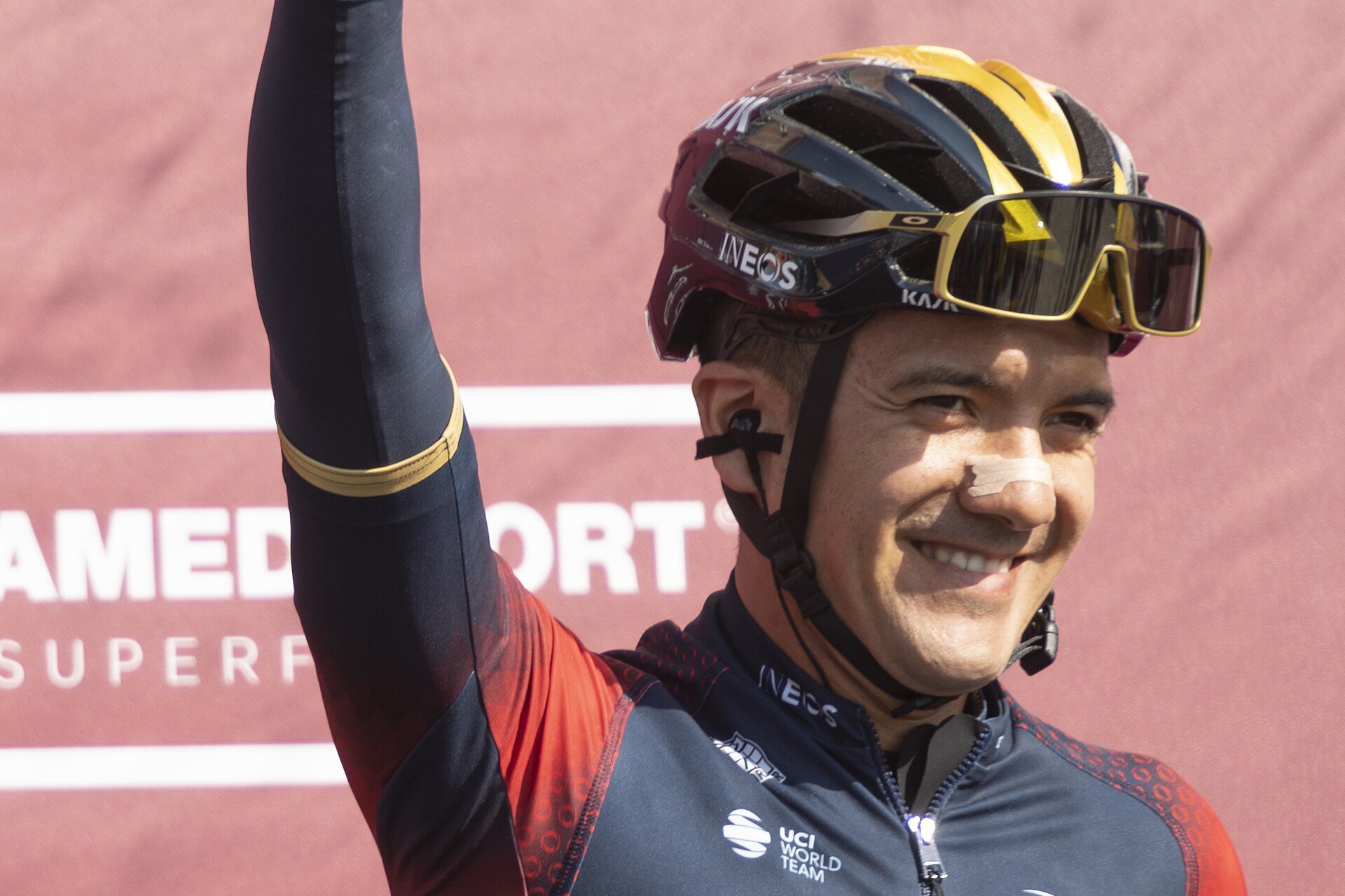 Cyclist Richard Carapaz smiles and raises an arm against a red backdrop, wearing an INEOS Grenadiers helmet and jersey