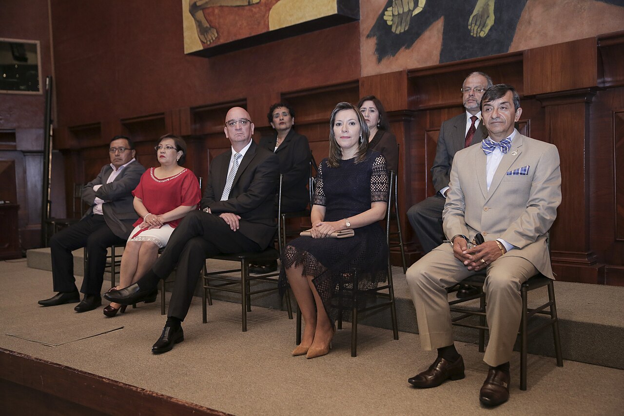Men and women in formal clothes sit on chairs on a stage in a wood-panelled hall beneath a large painted mural