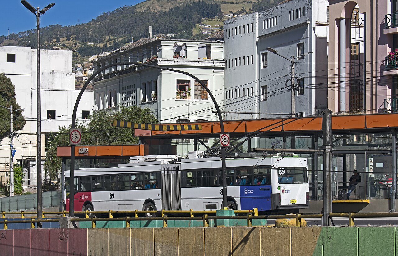 An articulated white trolleybus numbered 89 stands at a covered stop below old buildings and a green hillside in Quito