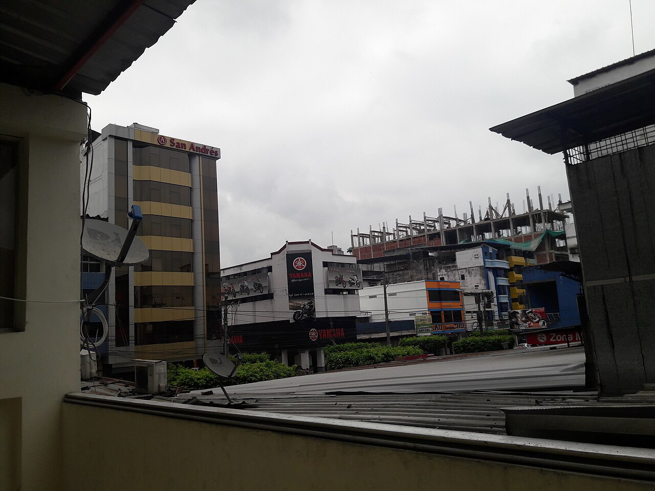 Commercial buildings in central Quevedo under a grey sky, including a gold and glass office tower and a motorcycle dealership