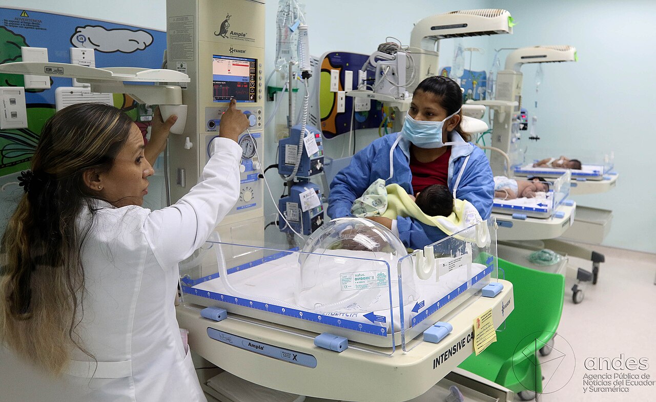 A doctor adjusts a monitor beside an incubator while a masked mother holds a baby in a hospital neonatal unit.