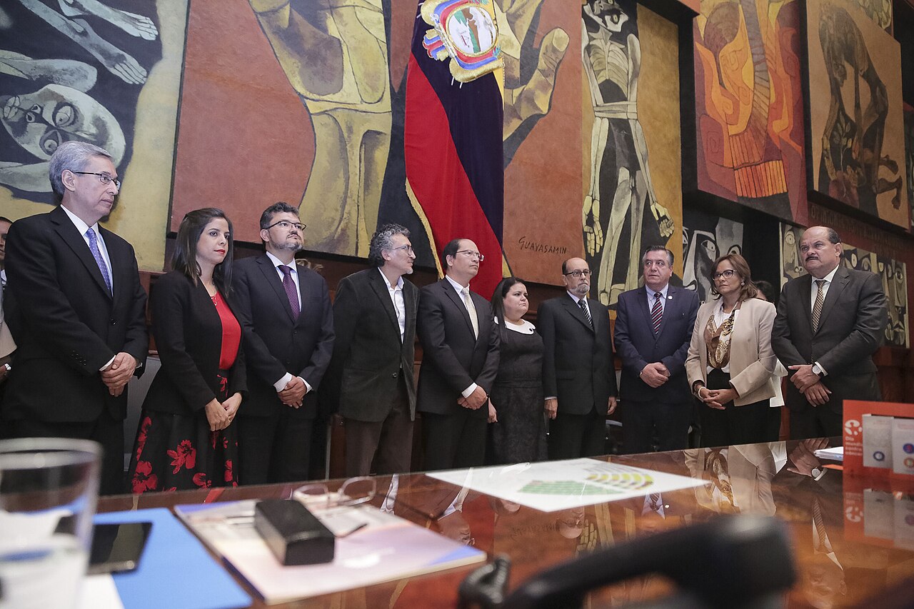 Judges and officials standing in the National Assembly chamber in Quito in front of a Guayasamin mural and the Ecuadorian flag
