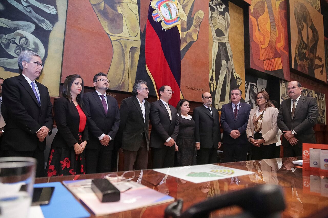 Judges and officials standing in the National Assembly chamber in Quito in front of a Guayasamín mural and the Ecuadorian flag