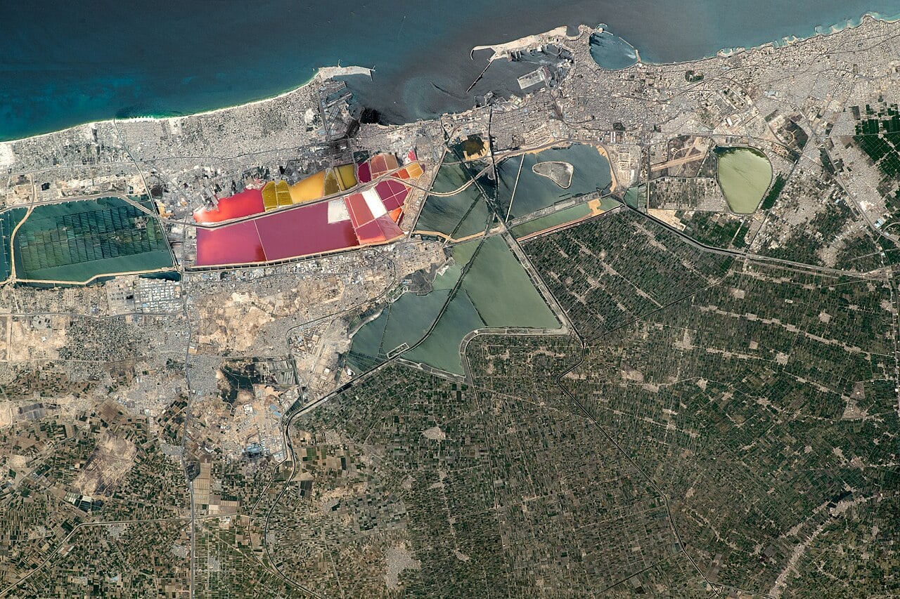 Astronaut photograph of Alexandria on Egypt's Mediterranean coast, with port breakwaters, a dense city strip, pink and yellow salt ponds, lagoons and green farmland inland