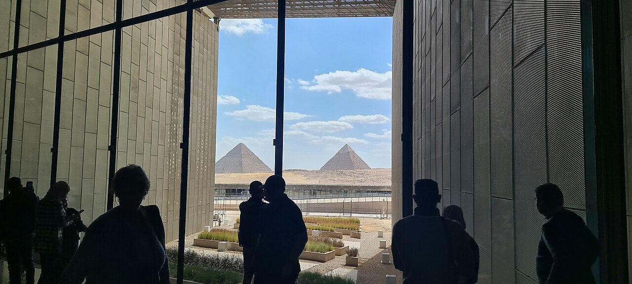 Visitors in silhouette look out from the Grand Egyptian Museum towards two Giza pyramids under a blue sky