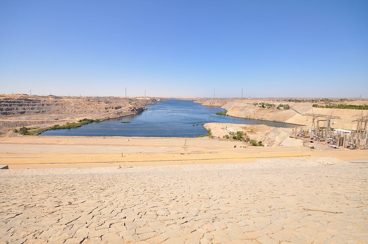The Nile flowing north between desert banks below the Aswan High Dam, with the dam's power station and pylons on the right