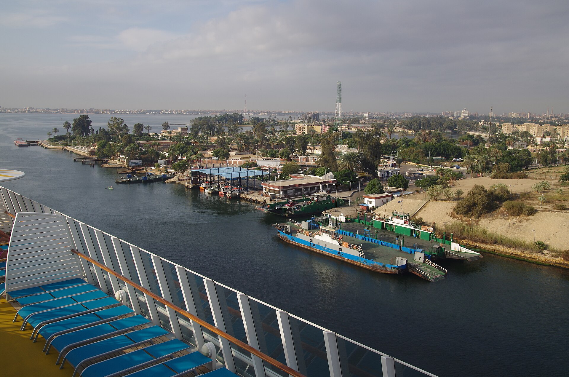 The Suez Canal at Ismailia seen from the deck of a passing cruise ship, with ferry pontoons, palm trees and the town behind