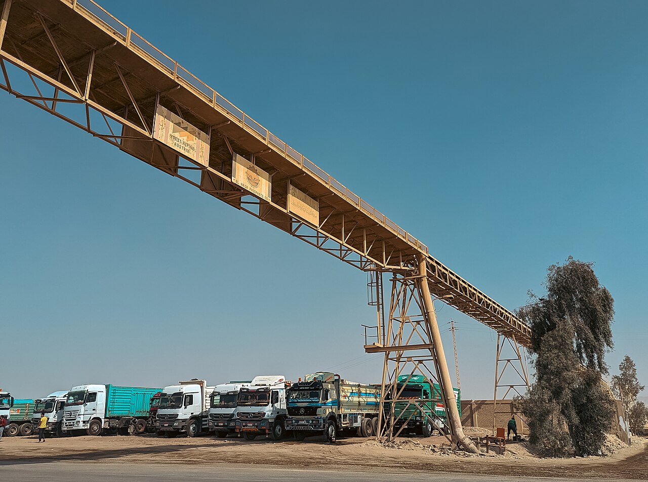 Lorries parked beneath a long conveyor belt bearing El Nasr Phosphate Company signs in the Egyptian desert