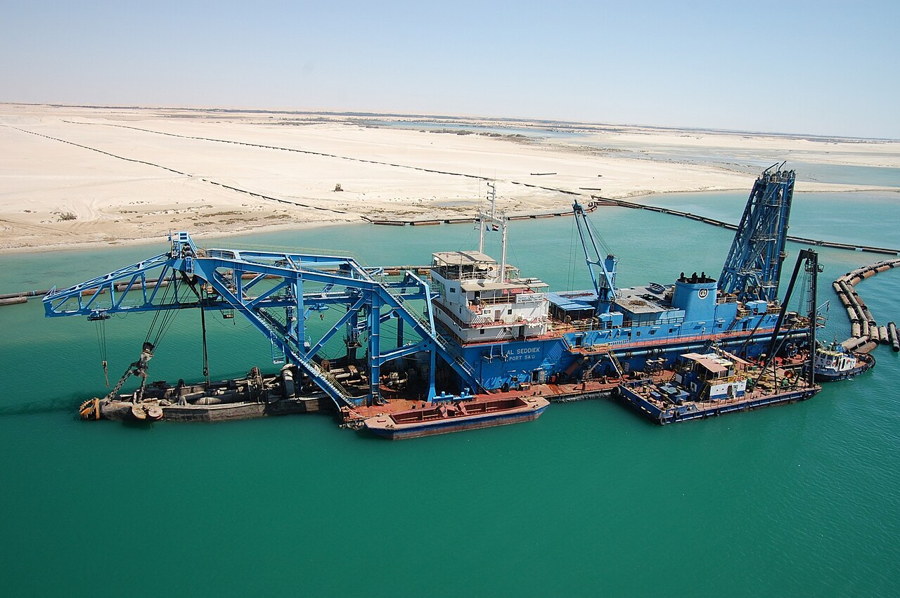 A blue dredger named Al Seddiek, registered in Port Said, working in the turquoise water of the Suez Canal beside desert sand banks