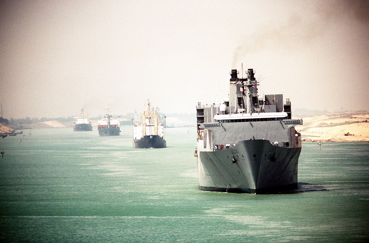 A grey US Navy cargo ship and three commercial ships move in line along the Suez Canal beside desert banks.
