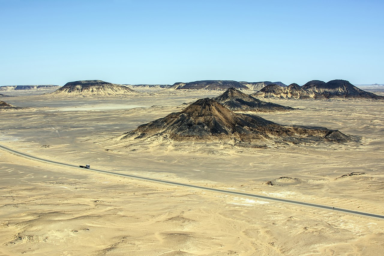 A paved road crosses a pale desert plain with dark flat-topped hills under a blue sky, with one lorry on the road