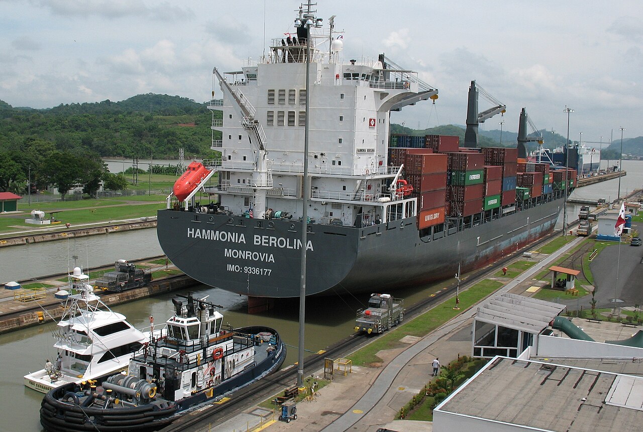 A container ship sits in a Panama Canal lock chamber, guided by a tugboat and electric towing locomotives on the lock walls