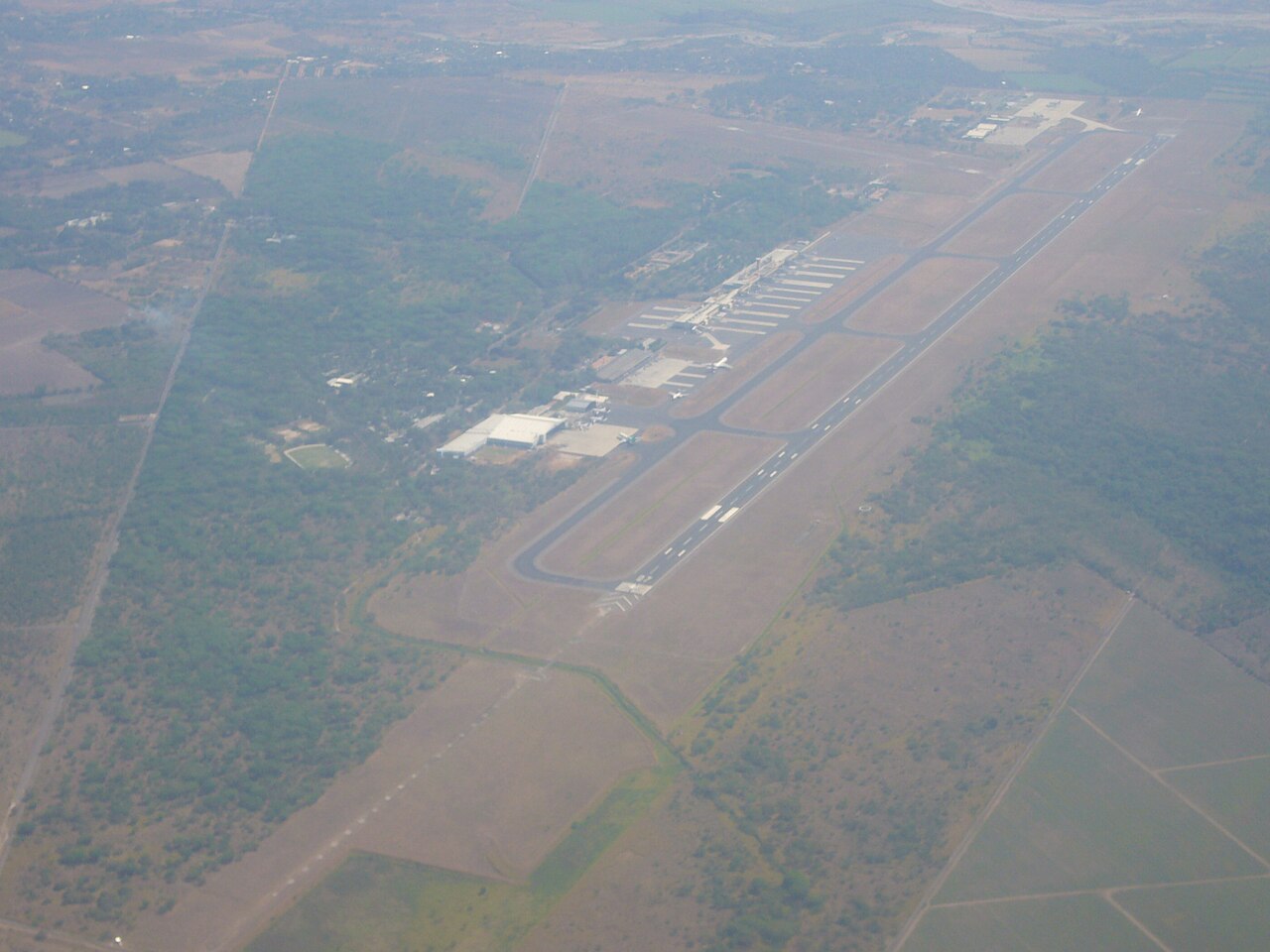 Aerial view of El Salvador International Airport with its terminal, apron and long runway surrounded by farmland and trees