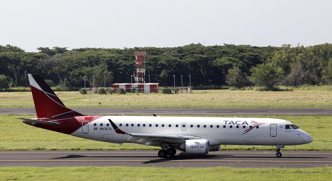 A red and white TACA Embraer passenger jet taxis along a runway at El Salvador International Airport