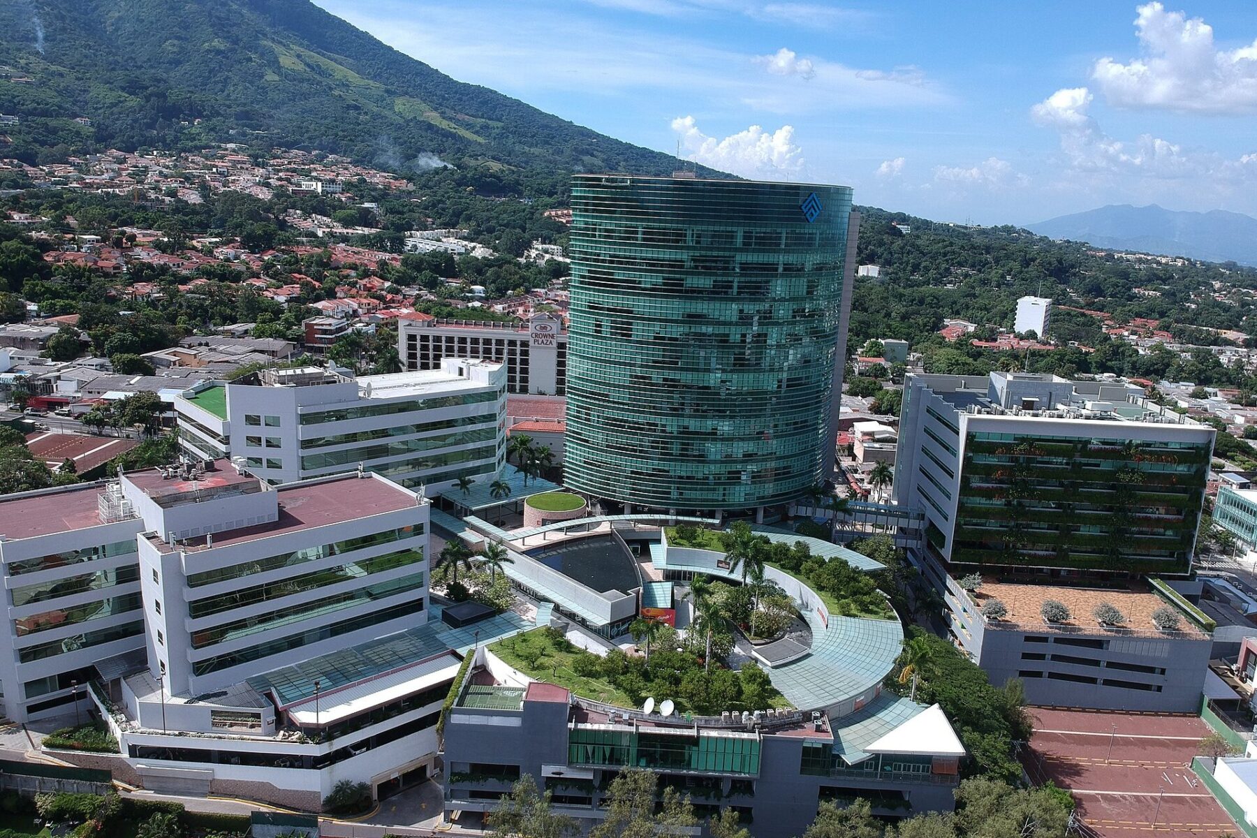 Aerial view of the glass tower and low office buildings of the World Trade Center San Salvador complex below a green mountain slope.
