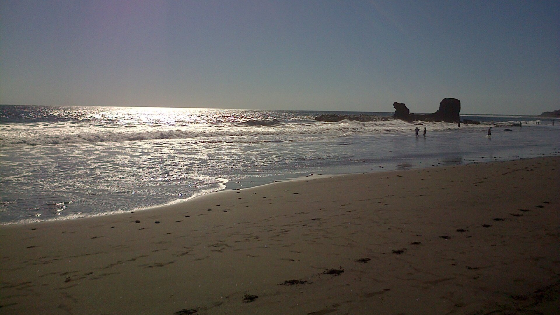 Waves break on the sandy shore of Playa El Tunco on the Pacific coast of El Salvador, with a dark rock and bathers in the distance.
