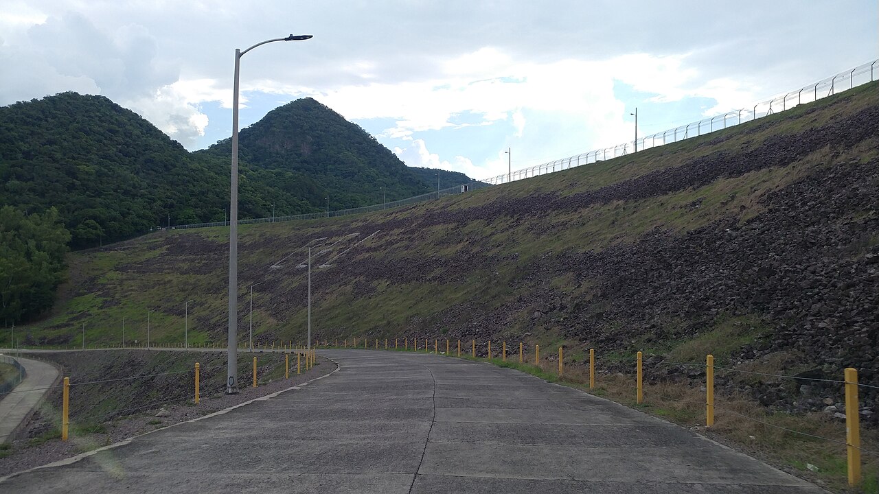 Road along the grass-covered rock embankment of the Cerrón Grande dam in El Salvador, with forested hills behind