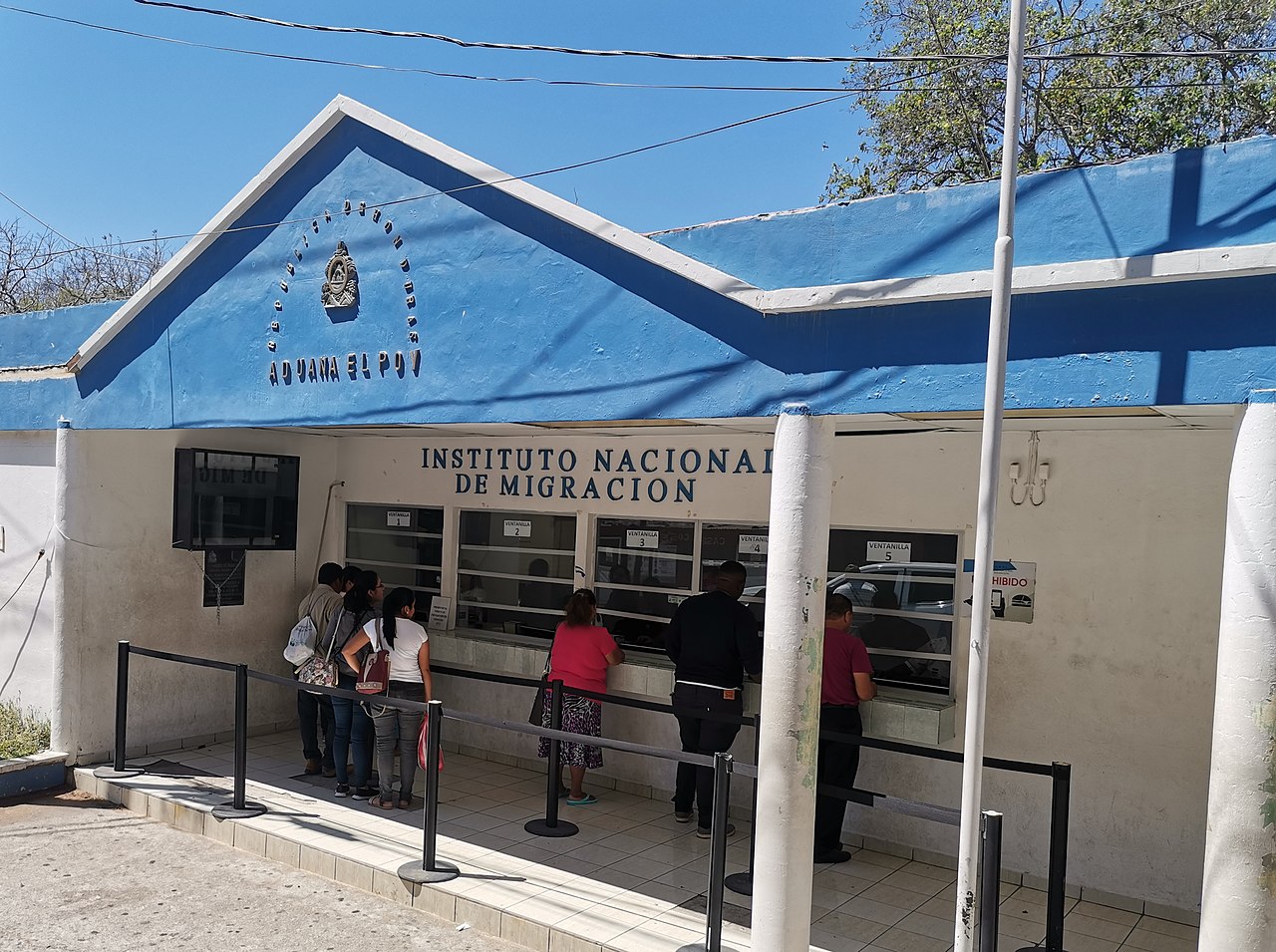 Travellers queue at the windows of a blue-and-white migration office marked Aduana El Poy and Instituto Nacional de Migración