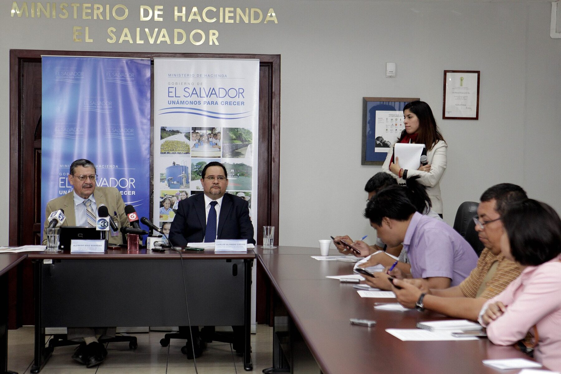Two officials seated at a table under a Ministerio de Hacienda El Salvador sign at a news conference, with reporters seated along the table at right