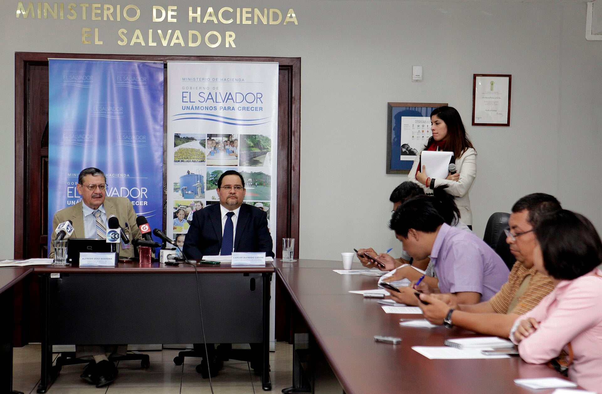 Two officials seated at a table under a Ministerio de Hacienda El Salvador sign at a news conference, with reporters seated along the table at right